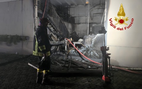 Vigili del fuoco in azione davanti a capannone in fiamme nella notte, squadre e mezzi di soccorso sul luogo, immagine di approfondimento